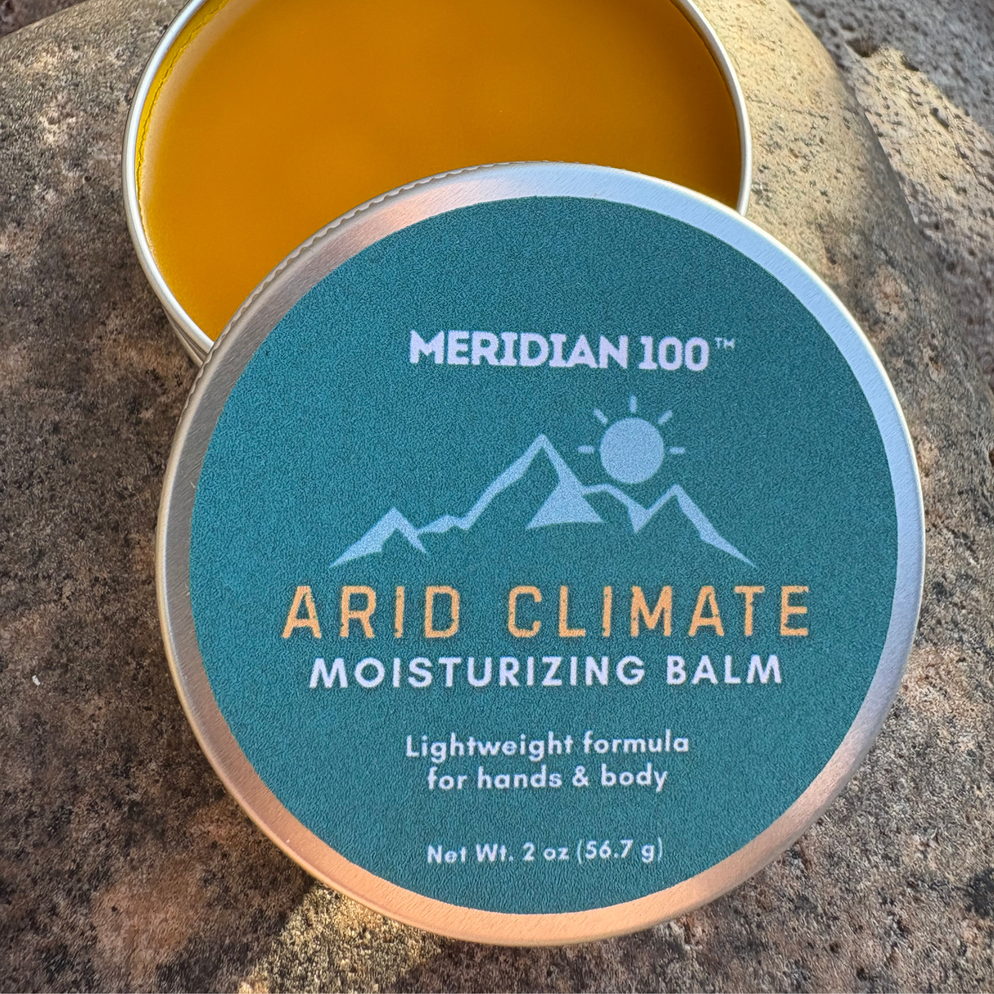 A round tin of Meridian 100 Arid Climate Moisturizing Balm, with the lid showing a teal label and the open tin showing a golden-yellow balm, set on a textured stone surface.