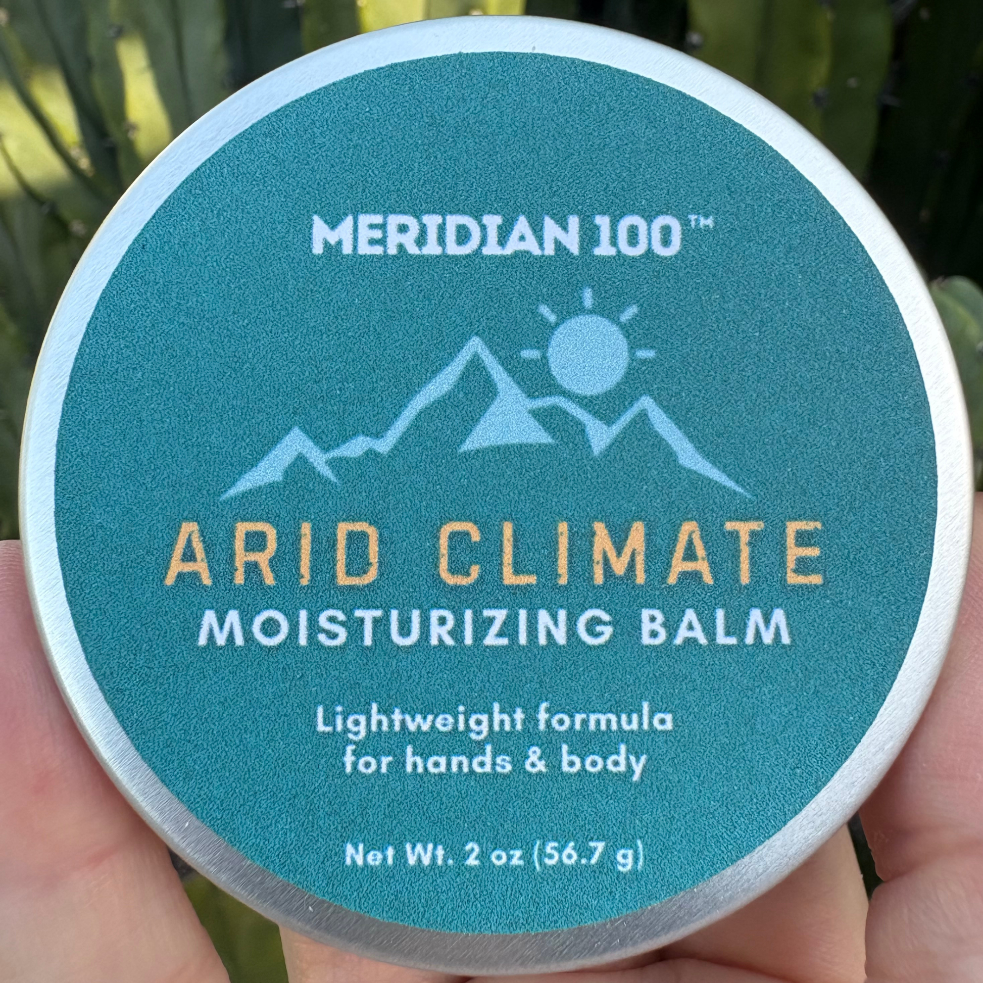A hand holding a round 2 oz sized tin of Meridian 100 Arid Climate Moisturizing Balm, showing the teal label with gold and white text against a blurred outdoor background.